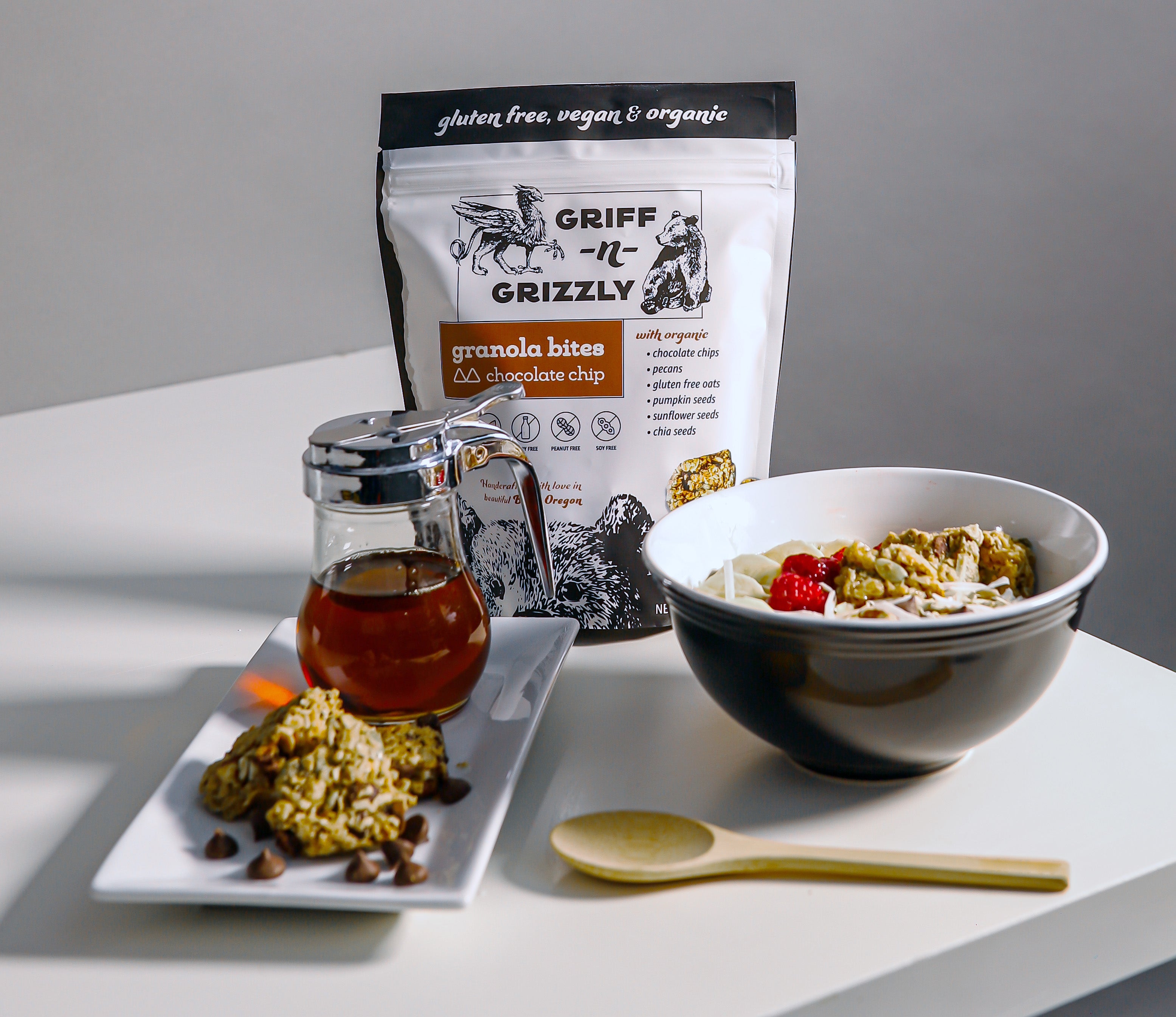 a plate with griff-n-grizzly granola bites and maple syrup next to a bowl of granola and fruit and a bag of granola bites in the background